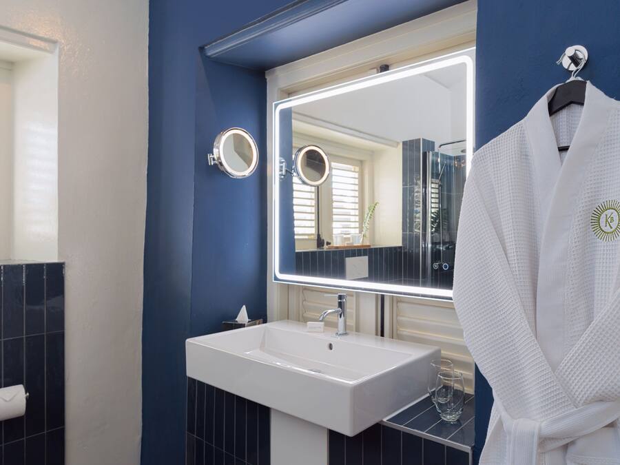 A bathroom with a white sink, a large mirror with lights, and a robe hanging on a rack.