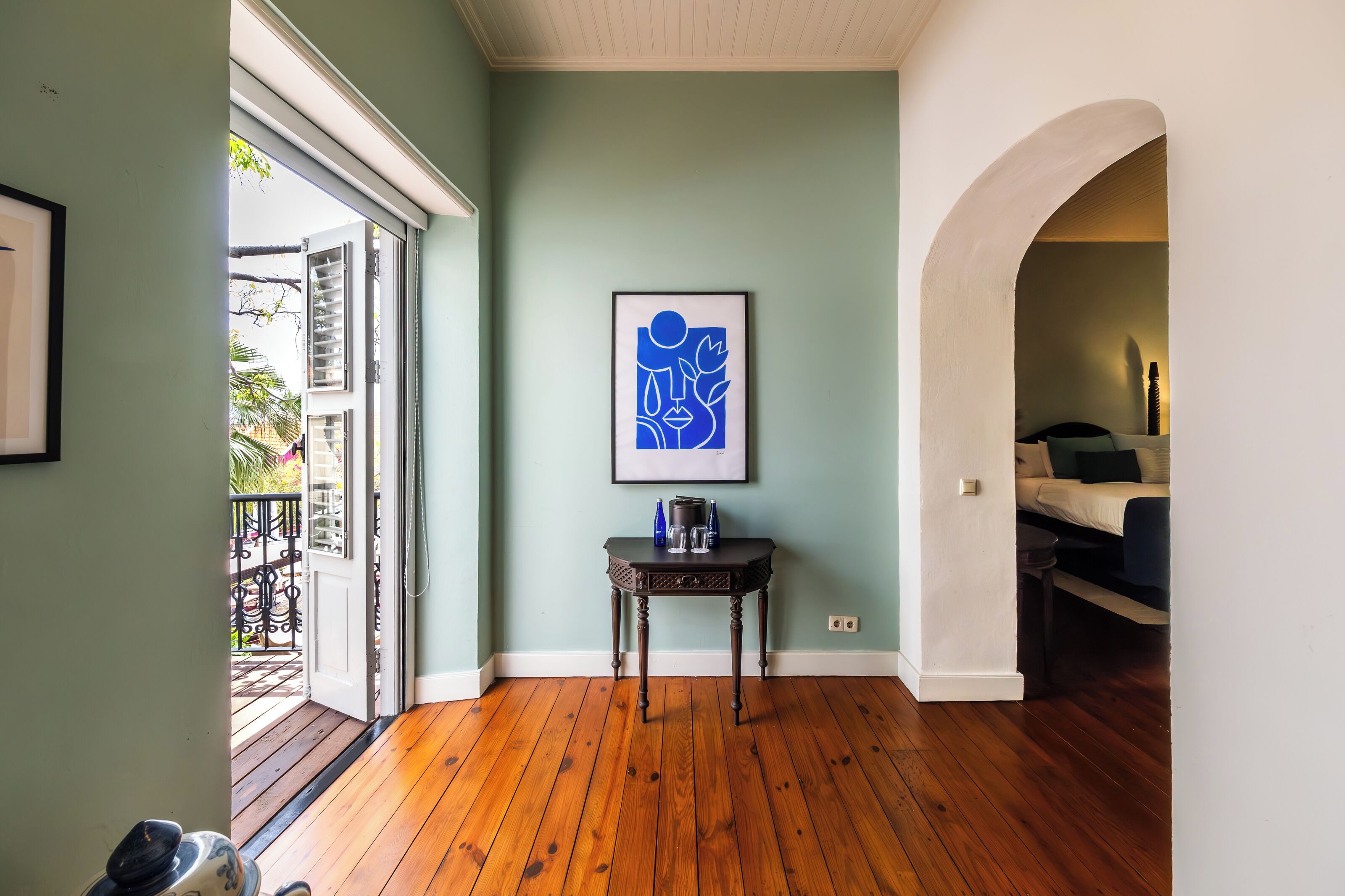 A room with wooden flooring, a framed abstract artwork on the wall, a small table with a coffee pot, and a view of a balcony with outdoor furniture.