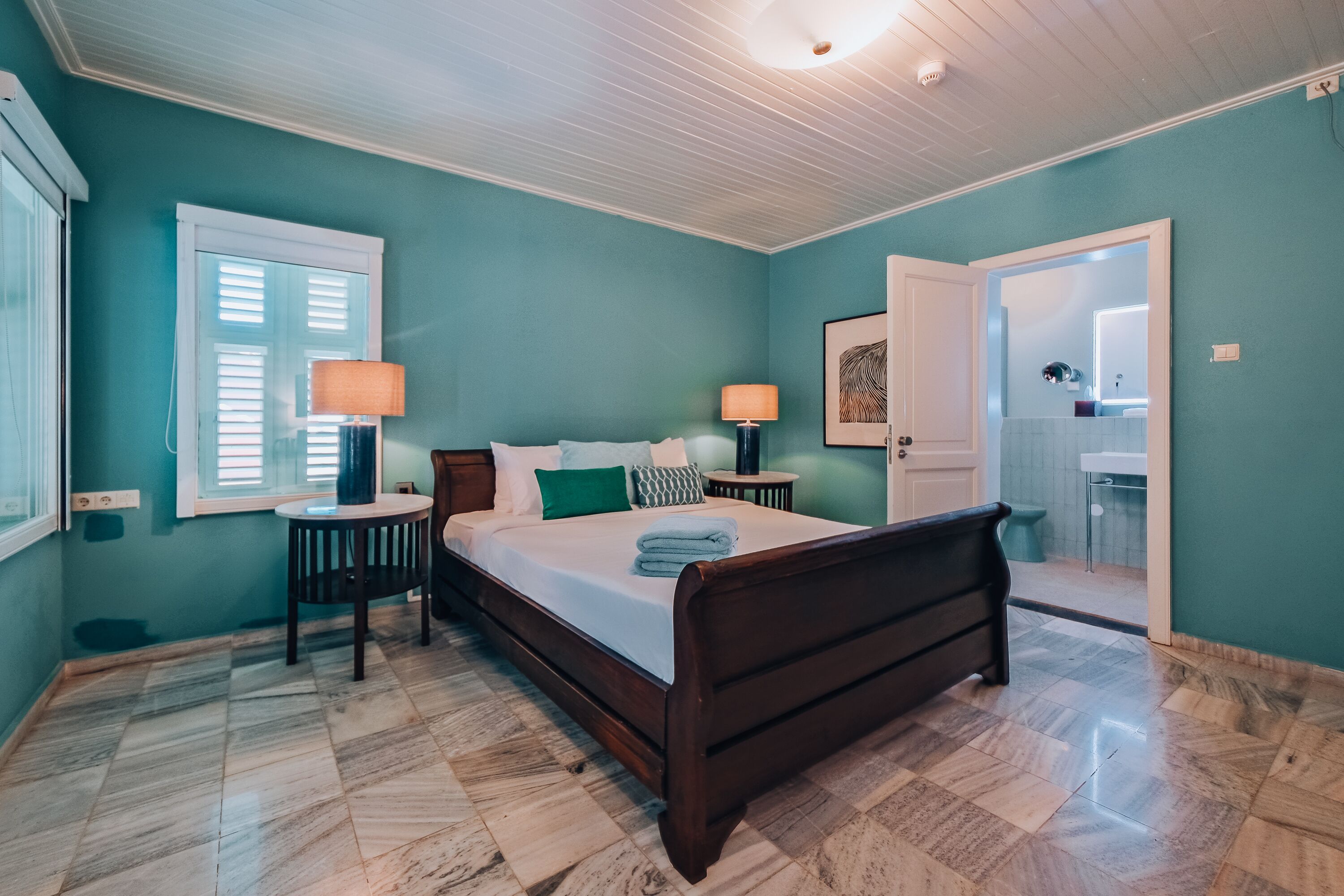 Un dormitorio con una cama de madera, dos mesitas de noche con lámparas, una ventana con persianas y una puerta que da a un baño.