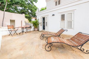 Un patio con sillones de madera y una mesa, un edificio blanco con contraventanas, y una planta en maceta.