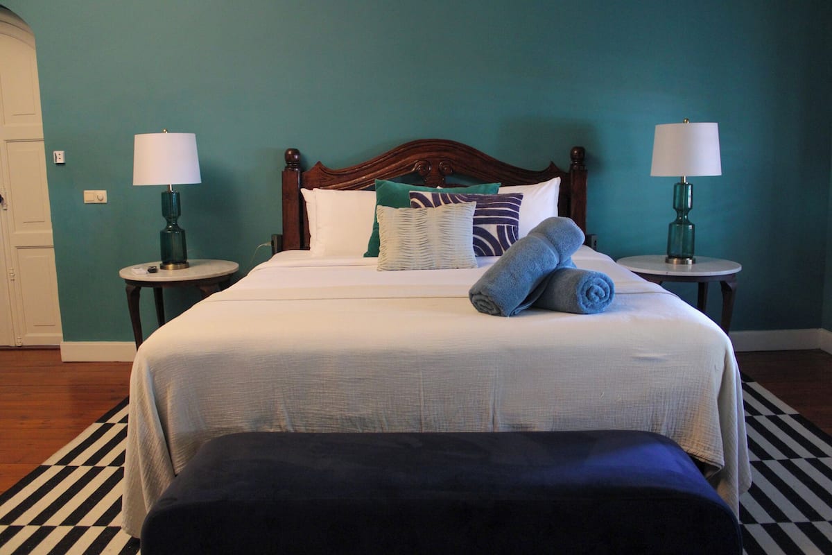 A neatly made bed with a wooden headboard, two bedside tables with lamps, a striped rug, and a bench at the foot of the bed.