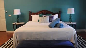 Una cama bien hecha con cabecera de madera, dos mesitas de noche con lámparas, una alfombra a rayas y un banco al pie de la cama.