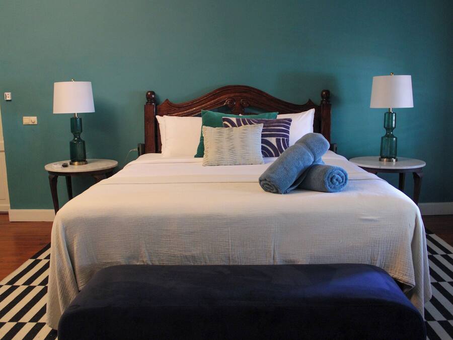 Una cama bien hecha con cabecera de madera, dos mesitas de noche con lámparas, una alfombra a rayas y un banco al pie de la cama.