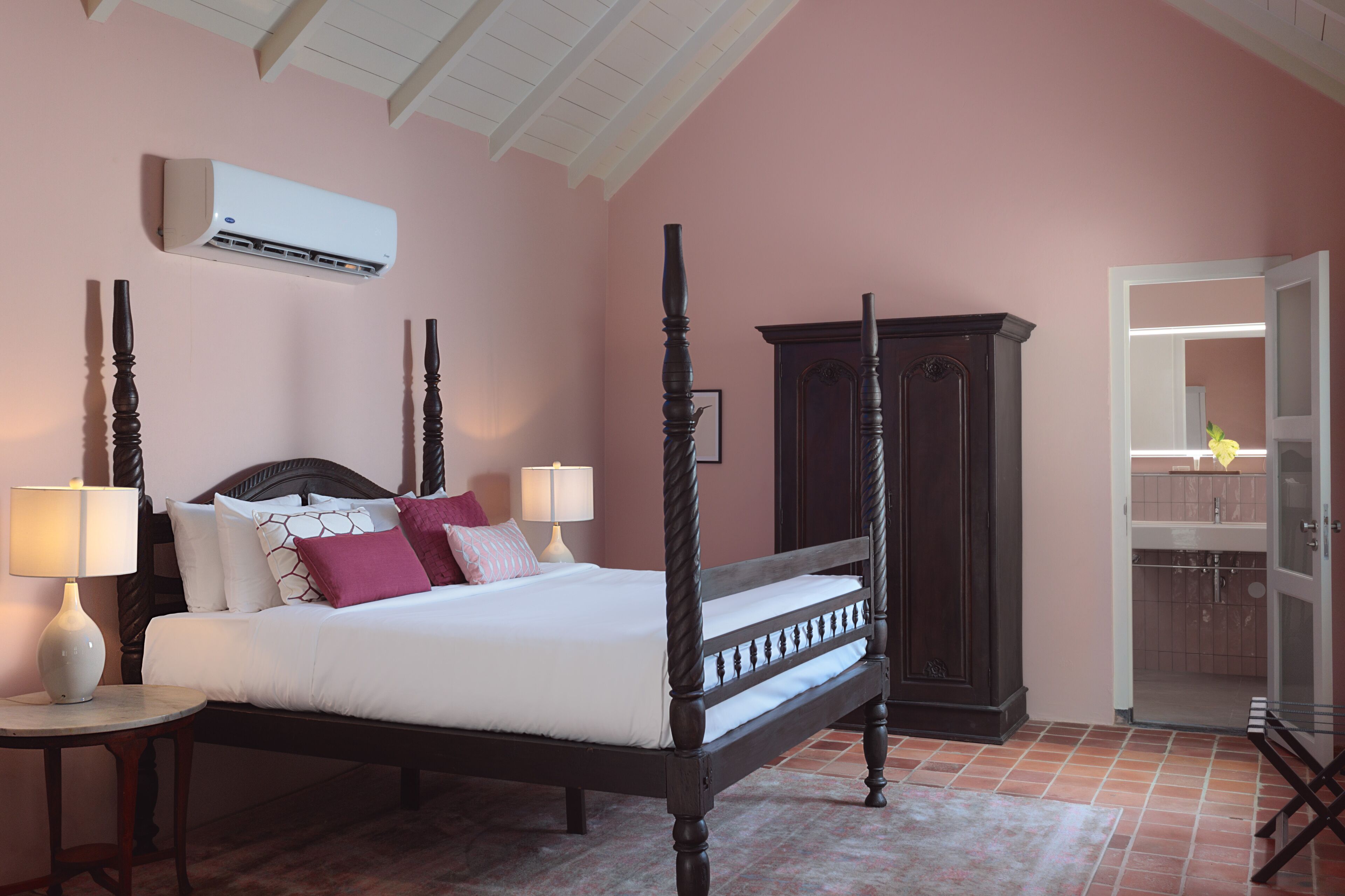 A bedroom with a four-poster bed, two bedside tables with lamps, a dark wooden wardrobe, and a bathroom visible through an open door.