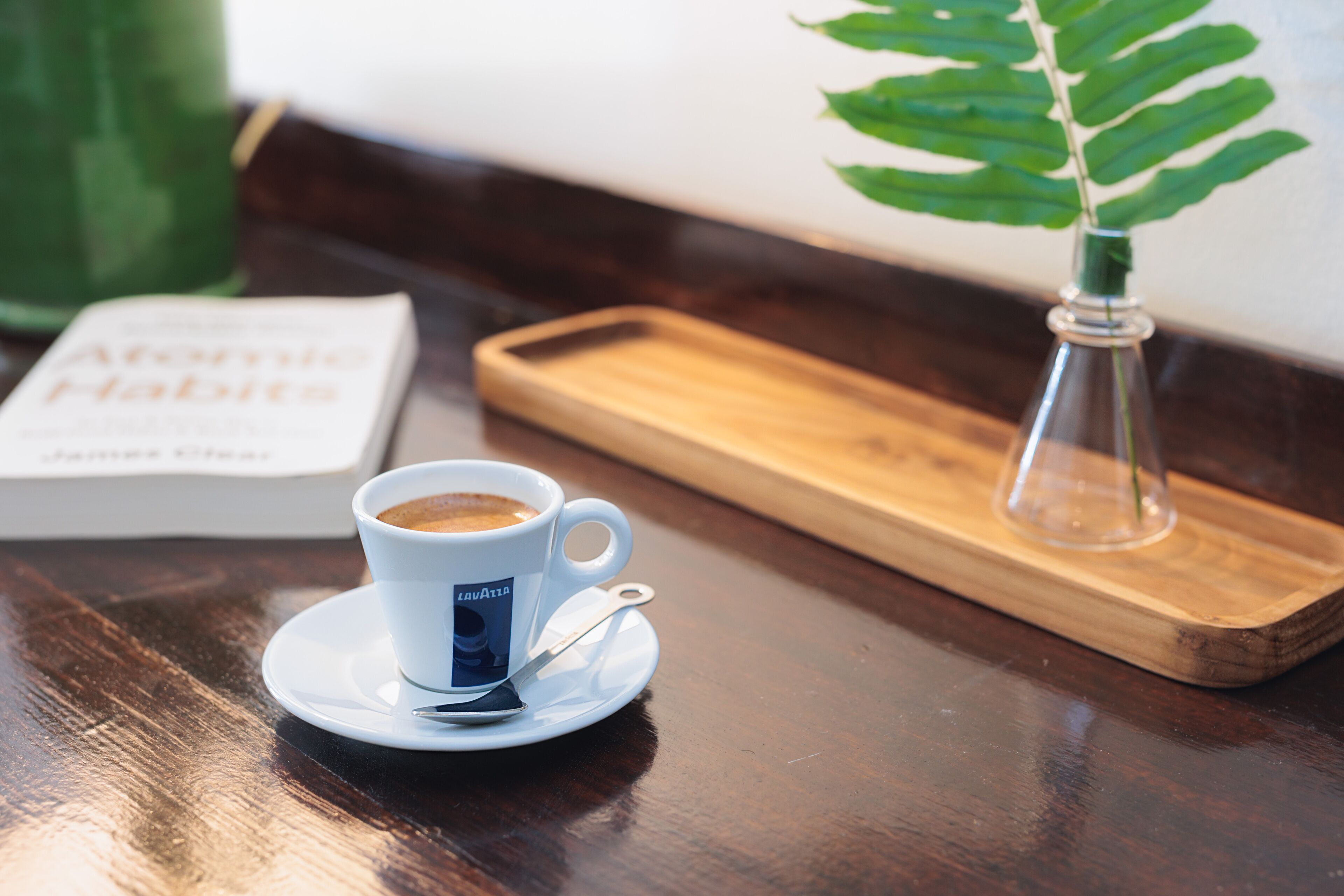 Una taza de café sobre un plato, un libro titulado “At Home Habits” sobre una bandeja de madera, y una pequeña planta en maceta sobre una mesa de madera.