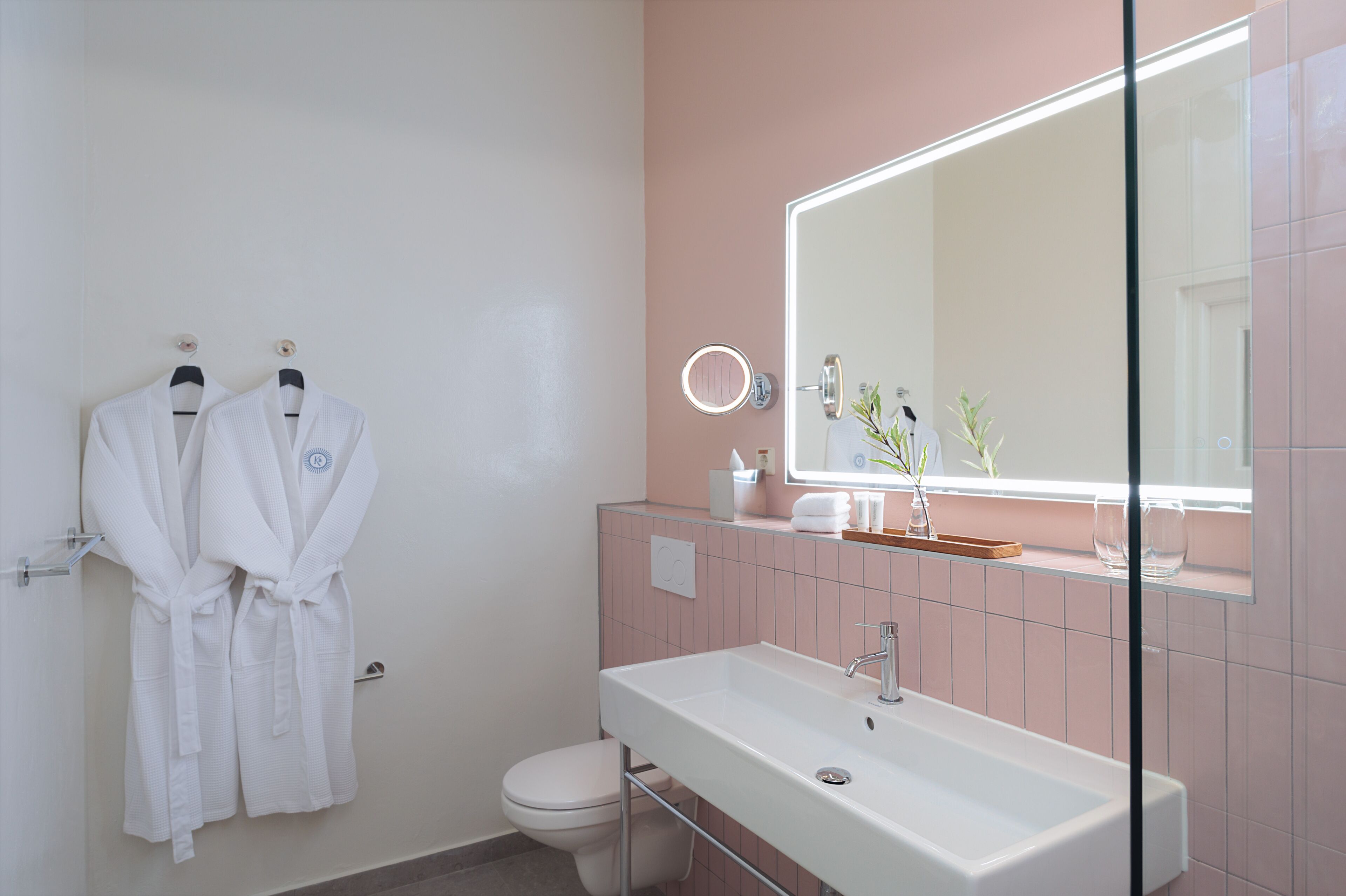 Un baño con un lavamanos blanco, un espejo y dos albornoces blancos colgados en la pared.