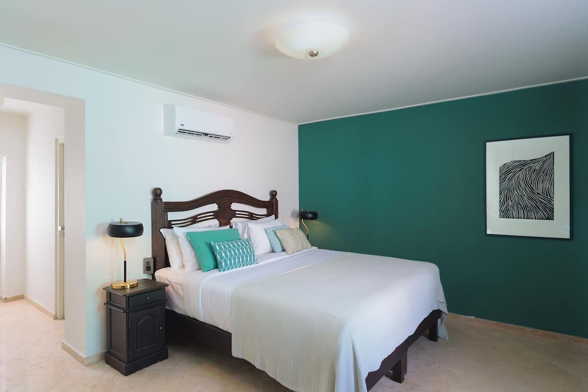 A bedroom with a bed, bedside tables, a nightstand, an air conditioning unit, and a framed artwork on the wall.