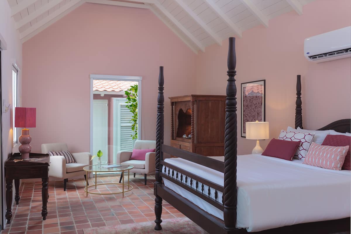 A bedroom with a four-poster bed, a sitting area with a chair and table, and a window with blinds.