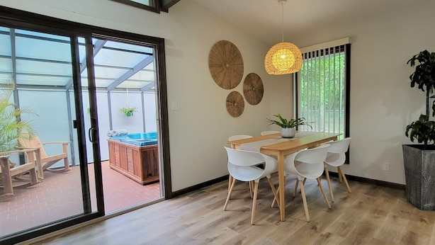 Uma área de refeições com mesa de madeira e cadeiras brancas, uma grande planta em vaso e uma porta de vidro deslizante que dá para uma área de estar ao ar livre.