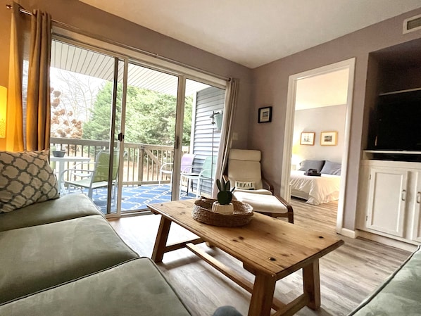 A cozy living room with a sofa, a wooden coffee table, and a chair. There is a sliding glass door leading to an outdoor area with a deck. - First floor 2br 2ba, balcony, pool, grill, pavilion, 1 block from Rehoboth Ave (Rehoboth Beach)