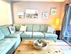 Una sala de estar con un sofá verde claro, una mesa de centro de madera con una cesta trenzada y tres obras de arte enmarcadas en la pared.