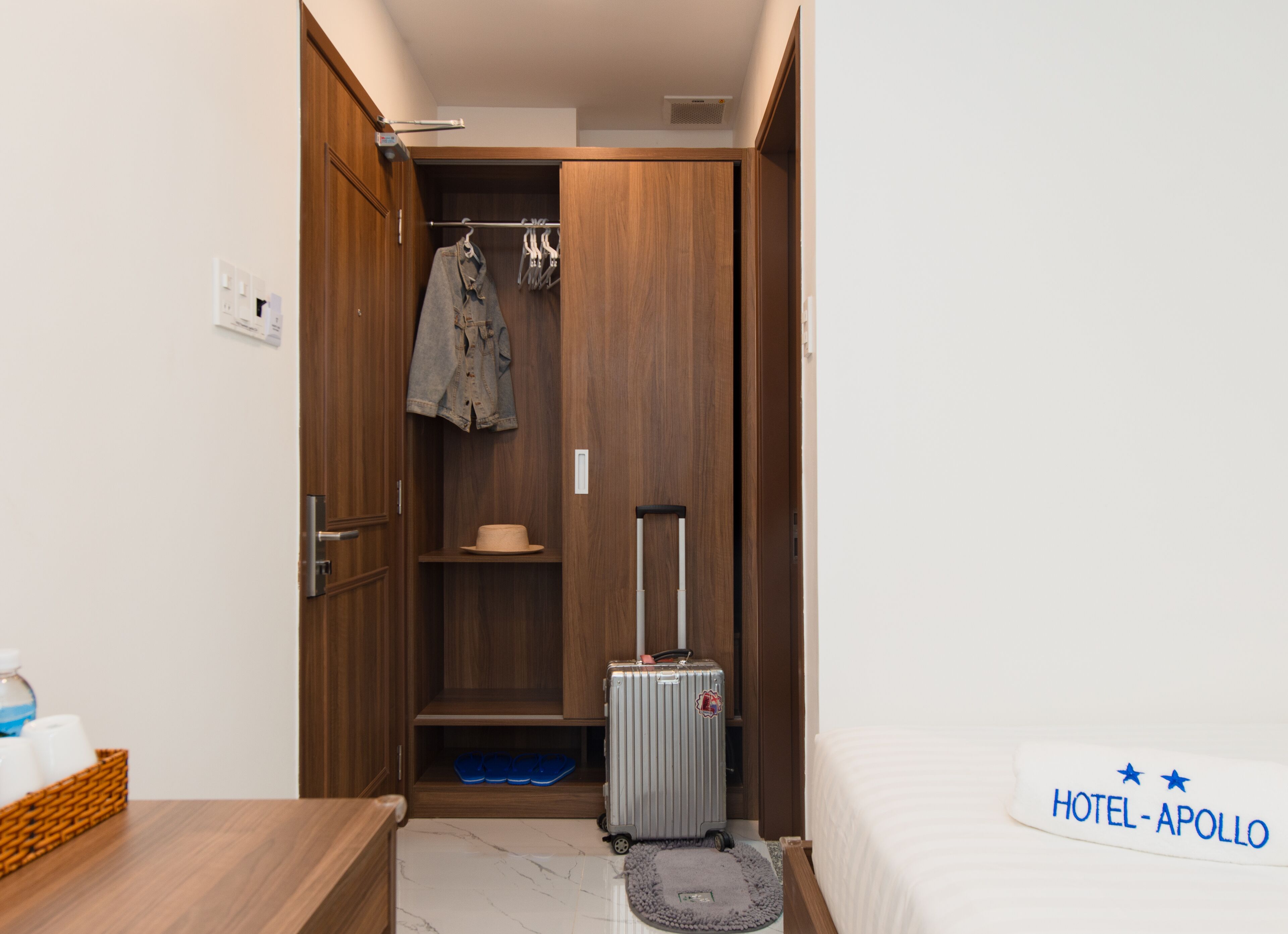 A compact hotel room with a wooden wardrobe, a suitcase, a hat, and a bed.