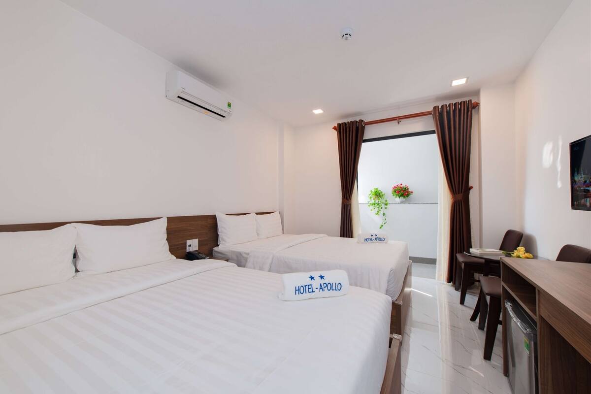 Quarto de hotel com duas camas, uma escrivaninha e varanda com cortinas.