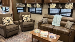 A cozy RV interior with a brown leather sofa, two armchairs, a coffee table with travel magazines, and patterned curtains. - Private Luxury Camping Experience  (Asheville)