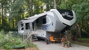 A large RV with a covered front area, a ladder, and a small porch with chairs and potted plants. - Private Luxury Camping Experience  (Asheville)