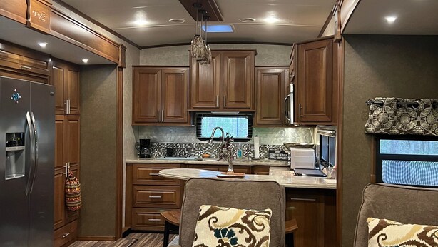 A compact RV kitchen with wooden cabinets, a refrigerator, a ceiling fan, and a dining area with two chairs. - Private Luxury Camping Experience  (Asheville)