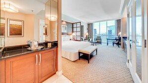 A hotel room with a large bed, a desk, a chair, a small table, a bench, a large window with a city view, and a kitchen area with a countertop and cabinets. - Jet Luxury Elite at The Signature (Las Vegas)