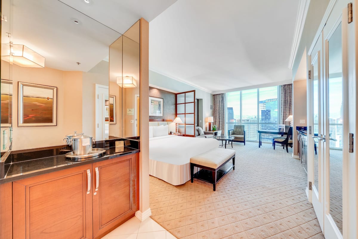 Hotelkamer met groot bed, bureau, stoel, klein tafeltje, bank, groot raam met stadszicht en een keuken met aanrecht en kastjes.