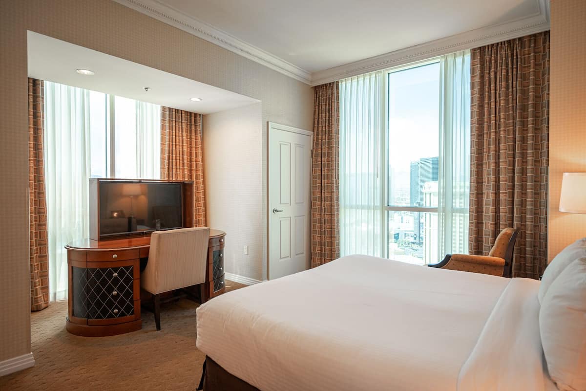 Een hotelkamer met een groot bed, een bureau met een televisie, en uitzicht op de stad door grote ramen.
