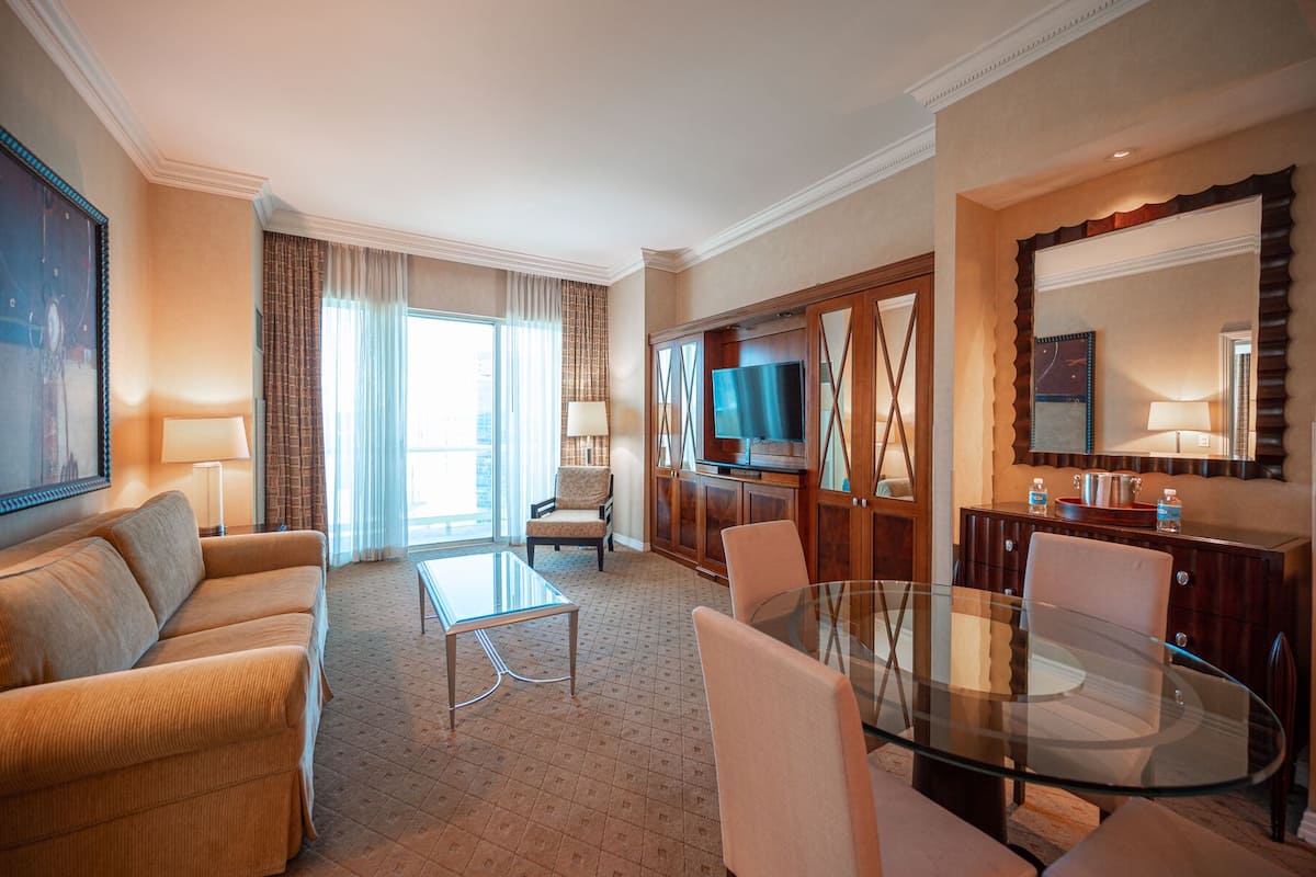 Een hotelkamer met een bank, een glazen salontafel, een televisie, een eettafel met stoelen, een grote spiegel en uitzicht op de stad door het raam.
