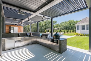 A modern outdoor kitchen with stainless steel appliances, a grill, and a view of a backyard with a pool and trees.