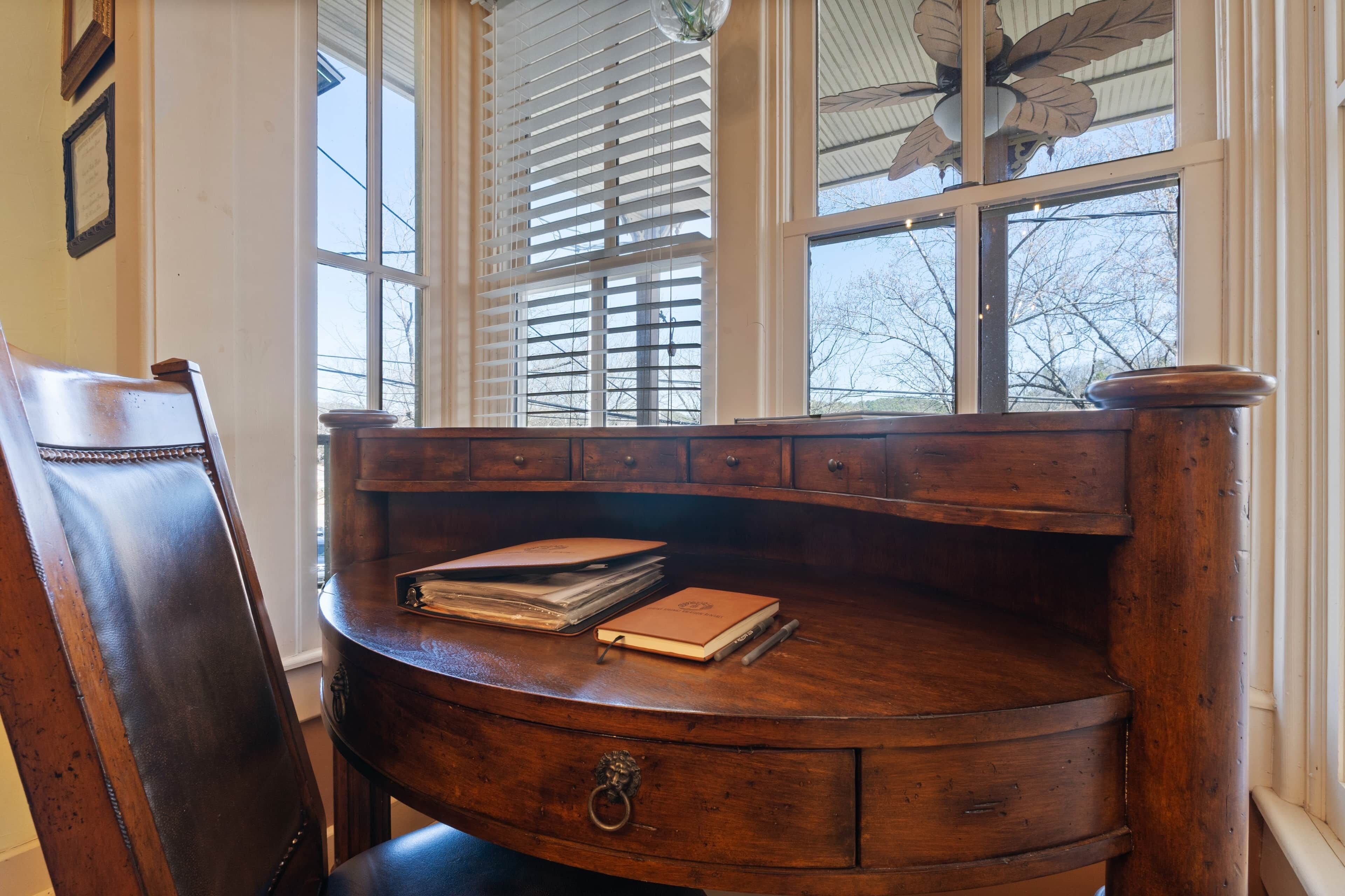 Un bureau en bois, une chaise en cuir, une fenêtre avec des stores et un ventilateur de plafond.