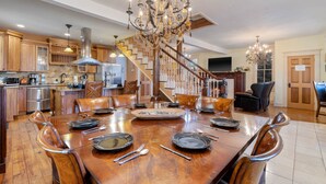 A spacious dining area with a large wooden table set for six, surrounded by wooden chairs. A grand chandelier hangs above, and a staircase with wooden railings leads to another level. - Downtown House w/Parking! On Spring St, Hot Tub! (Eureka Springs)