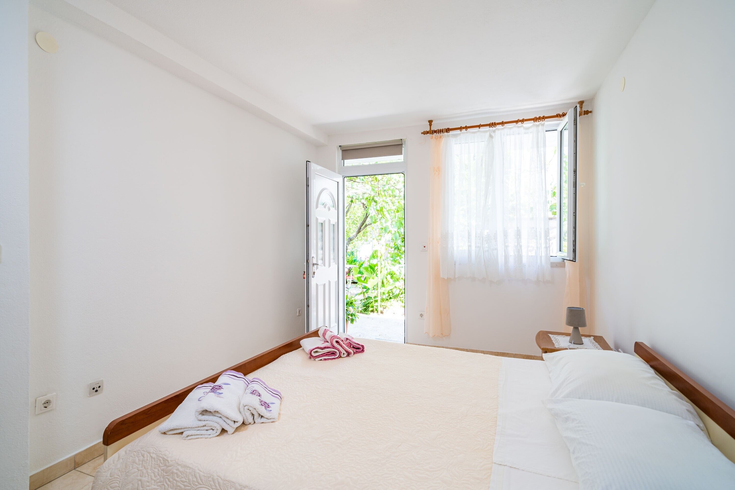 A bedroom with a bed, a nightstand, a window with curtains, and a view of greenery outside.