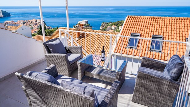 Eine Terrasse mit Korbmöbeln, einem Tisch und einer Weinflasche. Die Terrasse bietet Blick auf eine Küstenstadt mit orangen Dächern und das Meer.