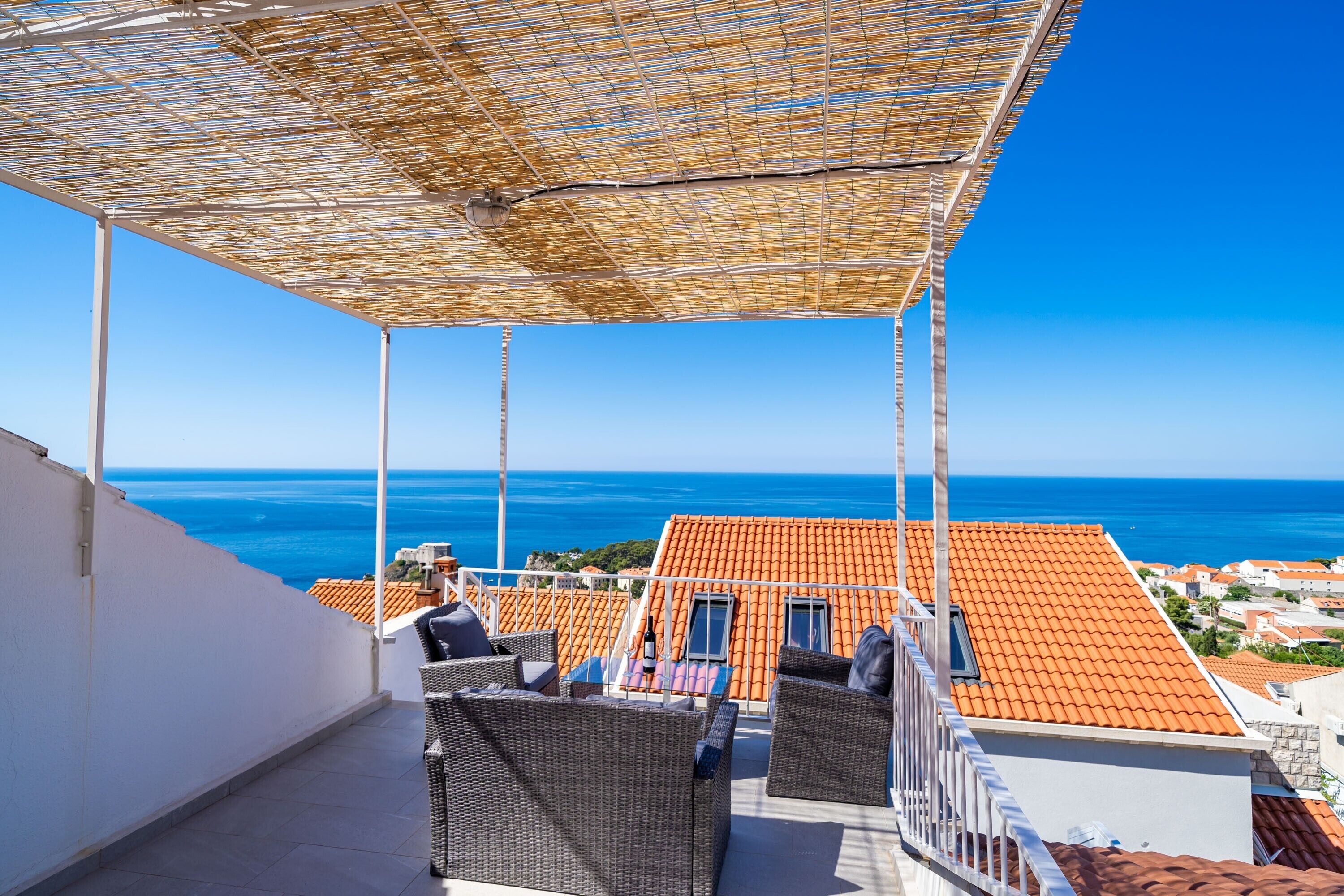 A balcony with wicker furniture, a view of the sea, and terracotta roofs.