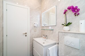 Un baño moderno con una puerta blanca, un lavabo con un espejo encima, un secador de pelo, un jarrón con flores moradas y un perchero para toallas.