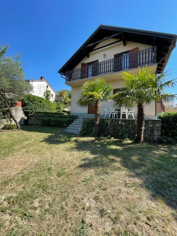 Une maison à deux étages avec un balcon, un mur en pierre et une cour gazonnée.