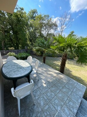 Eine Terrasse im Freien mit einem runden Tisch und weißen Stühlen, umgeben von einem steinbelagten Boden und üppiger Vegetation.