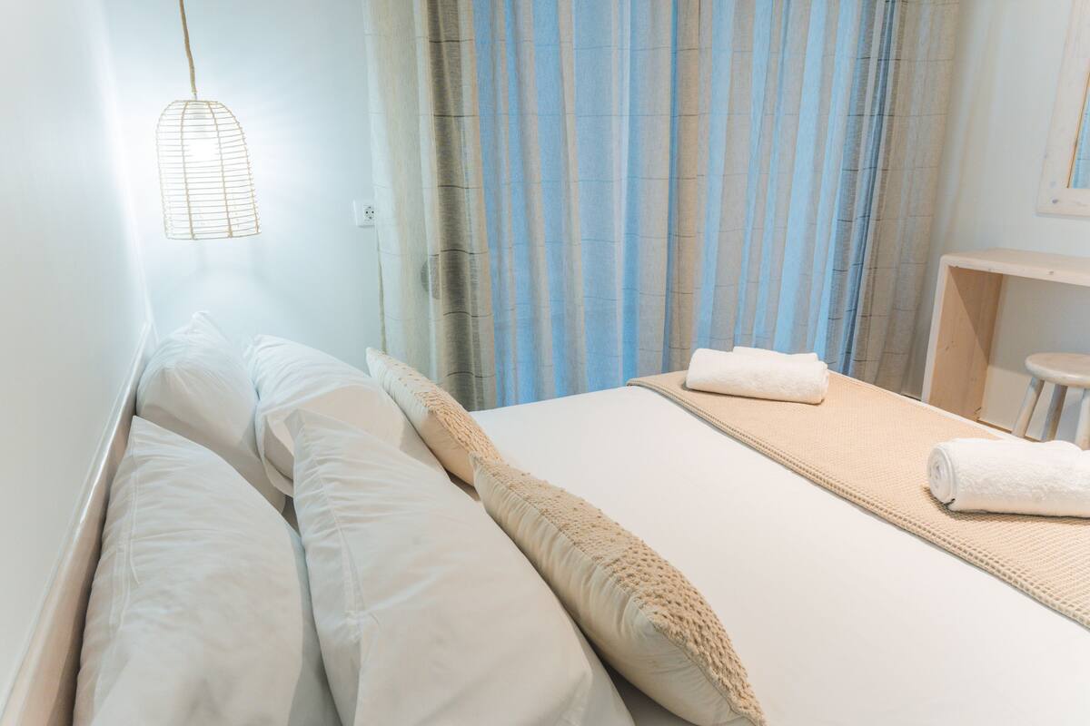 Een net opgemaakt bed met witte lakens en een beige plaid, een klein nachtkastje met een handdoek, en een hanglamp.