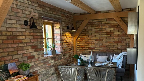 Une cuisine rustique avec des poutres apparentes, un mur en briques, un coin repas avec des chaises en osier et un petit meuble contenant un téléviseur.