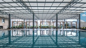 Una piscina interior con suelo de vidrio, rodeada de sillones de madera y una estructura de techo metálica.