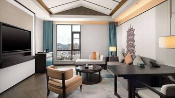 A modern hotel room with a large TV, a sofa, a dining table, and a view of the city.
