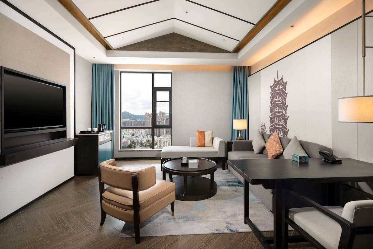 Habitación de hotel moderna con televisor grande, sofá, mesa de comedor y vistas a la ciudad.