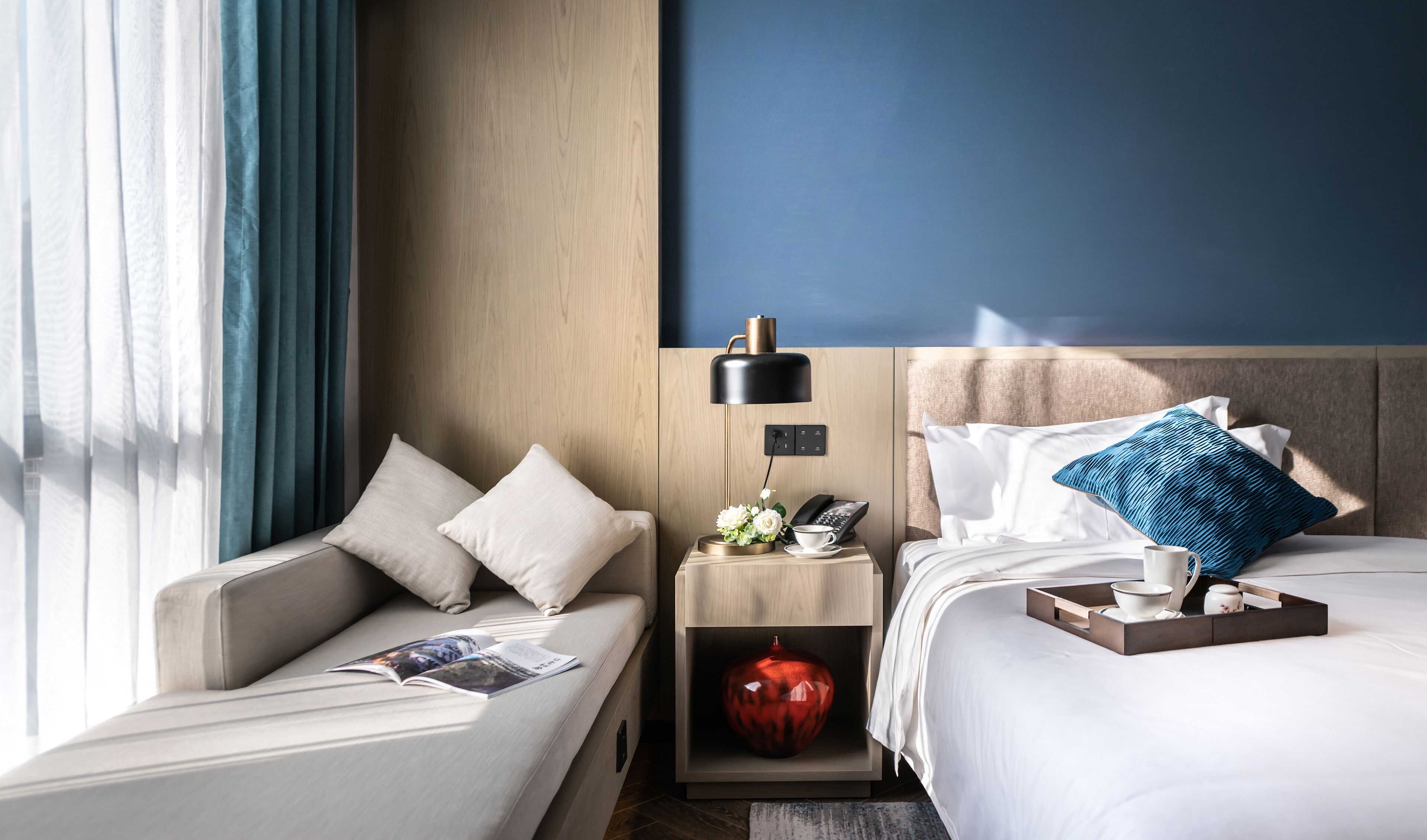 Een hotelkamer met een sofa, een nachtkastje met een lamp, een telefoon, een klein decoratief object en een bed met kussens en een dienblad.