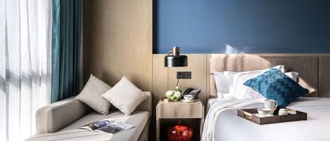 Een hotelkamer met een sofa, een nachtkastje met een lamp, een telefoon, een klein decoratief object en een bed met kussens en een dienblad.