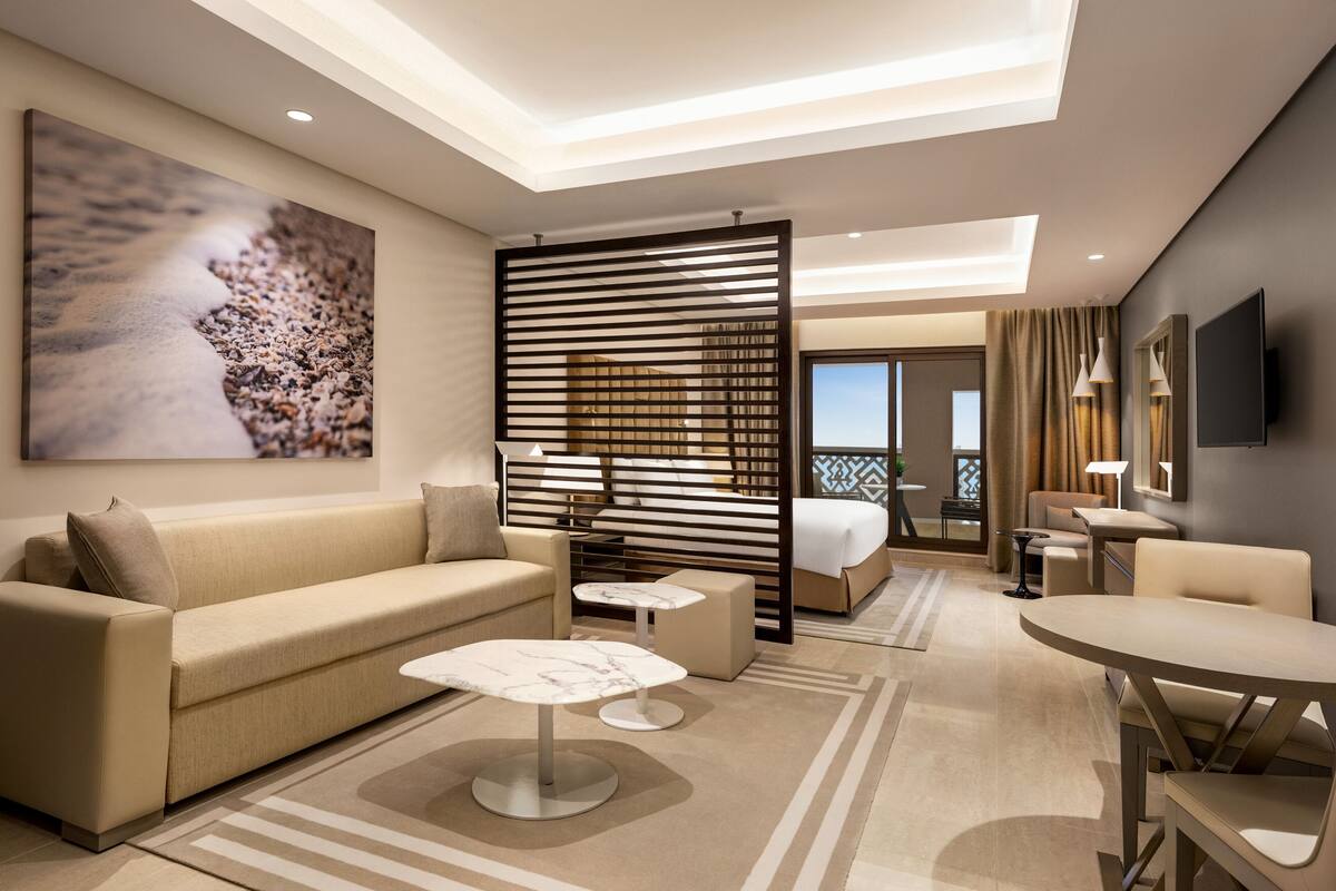 A modern hotel room with a sofa, a dining table, a coffee table, a large artwork, a sliding door, and a balcony view.