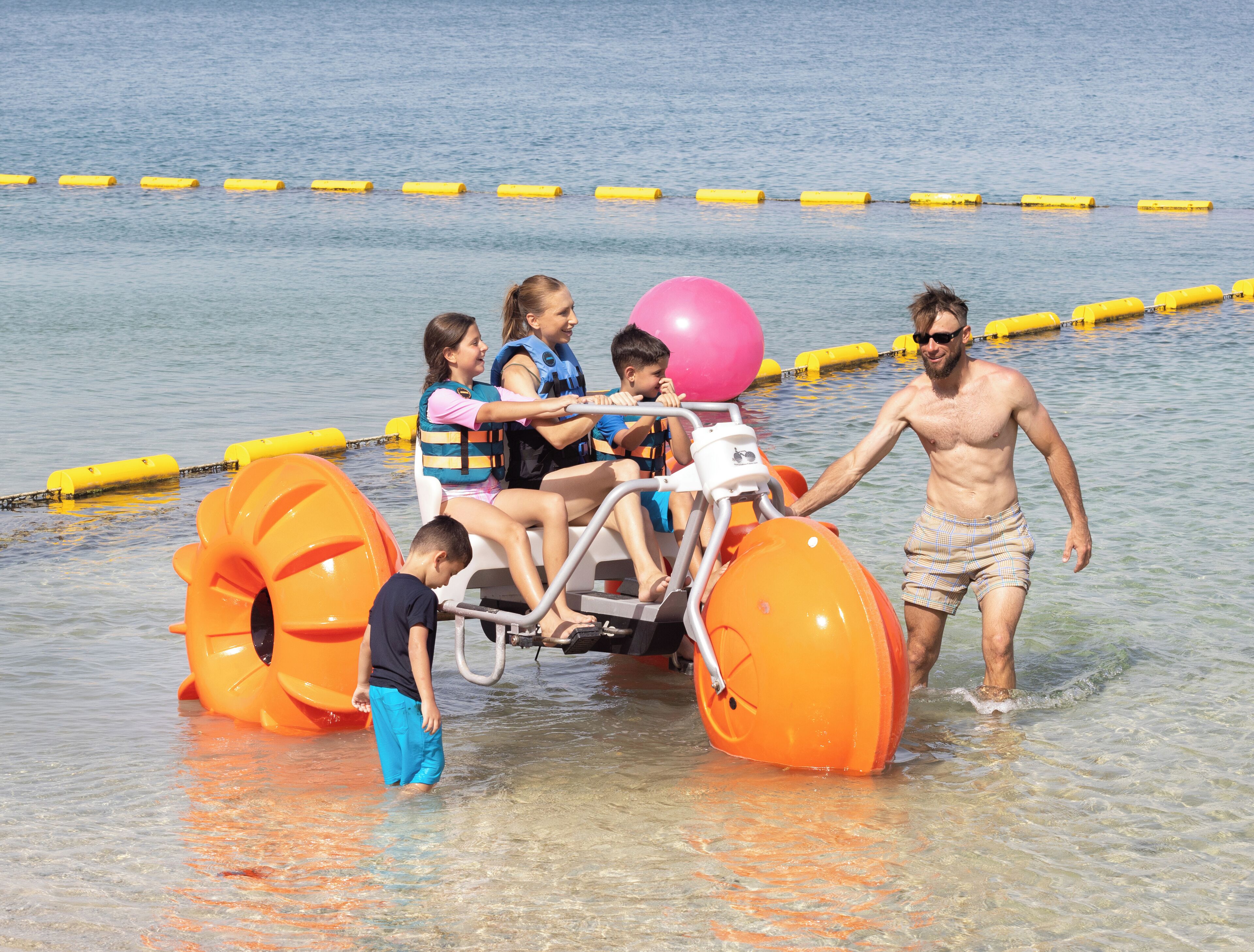 A water ride with a man pushing a vehicle carrying children and a pink balloon.