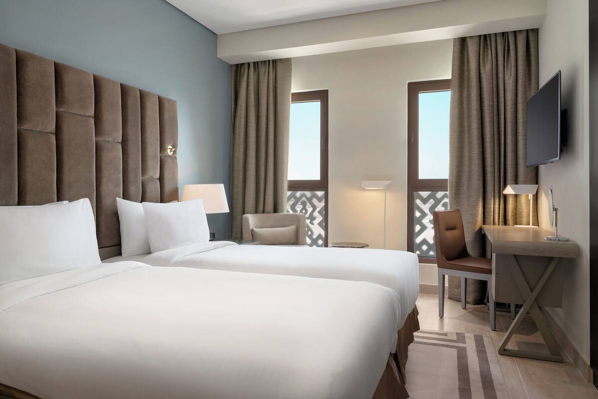 A hotel room with two beds, a desk, a chair, and a TV.