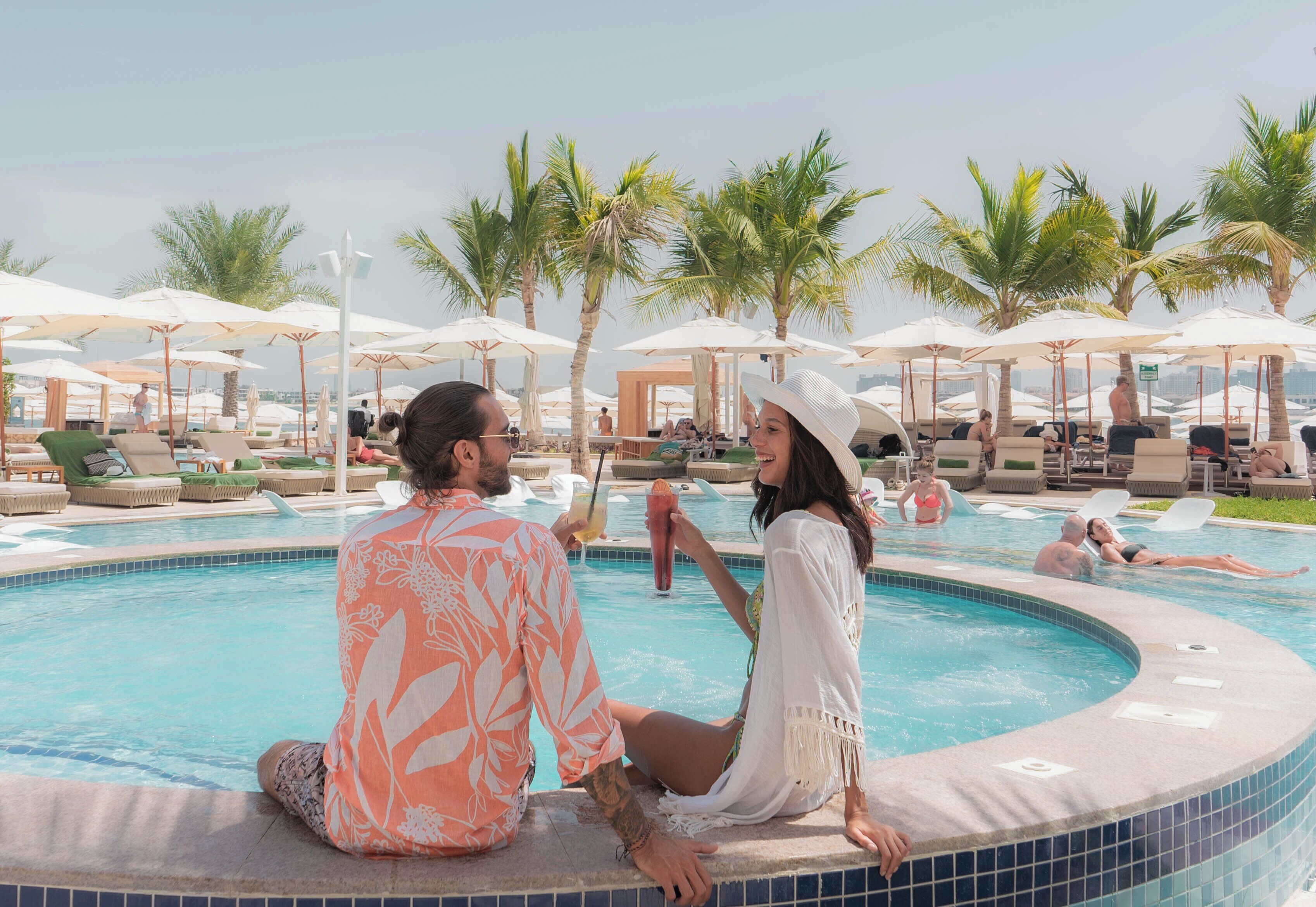 A poolside area with lounge chairs and umbrellas, palm trees, and a clear sky.