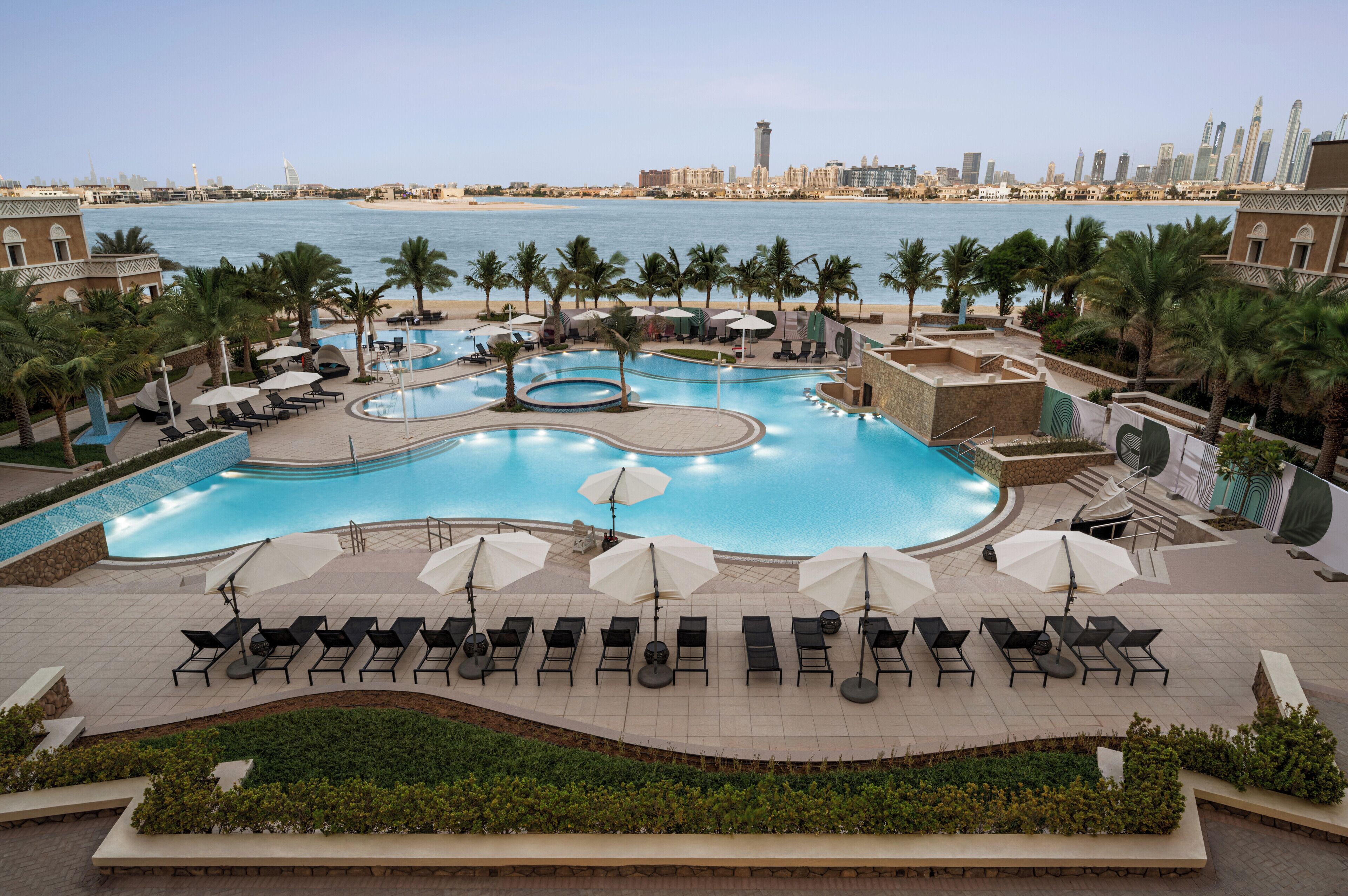 A resort pool area with multiple sun loungers and umbrellas, surrounded by palm trees and a city skyline in the background.
