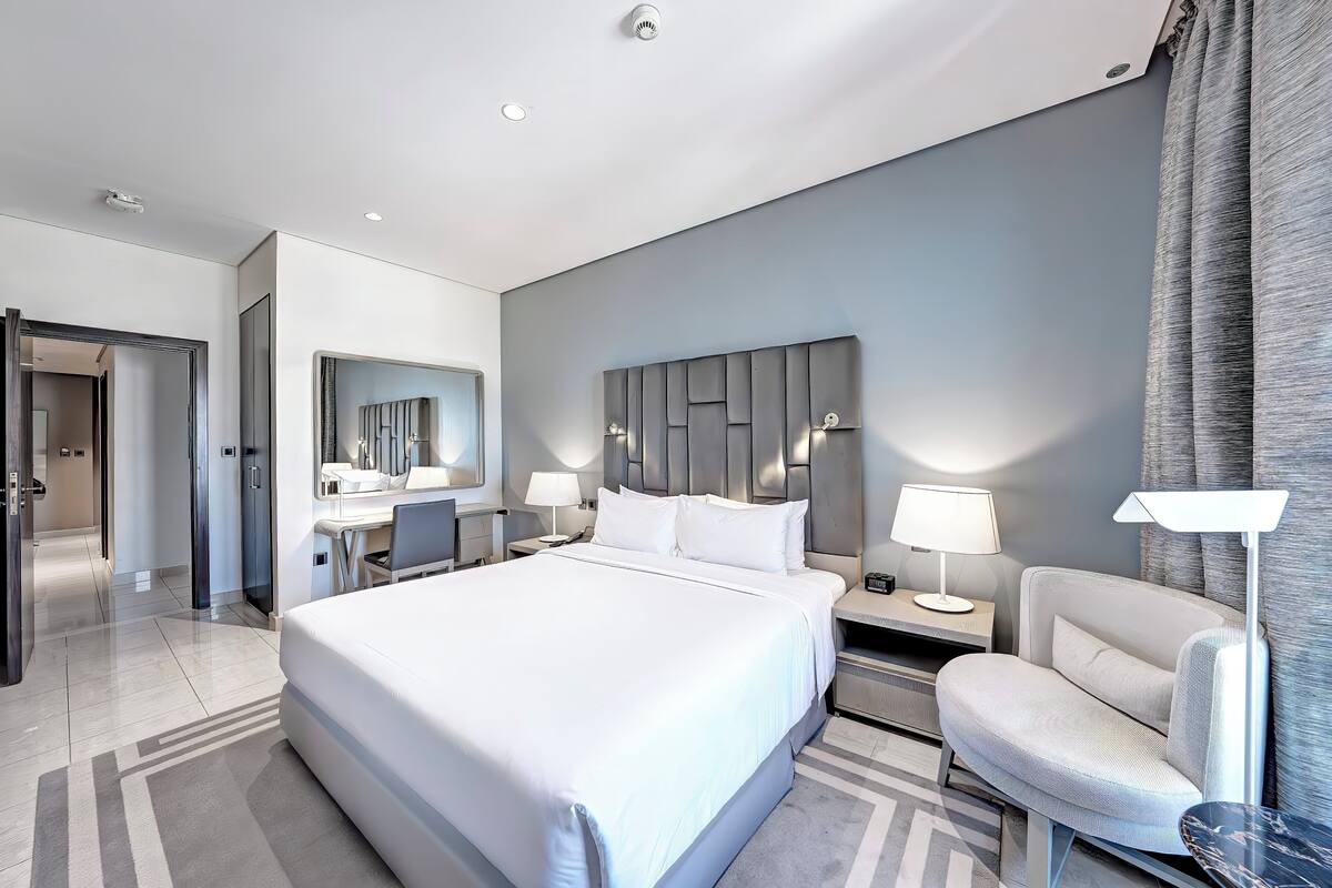 A modern hotel room with a large bed, a desk with a chair, a vanity with a mirror, and two bedside lamps.