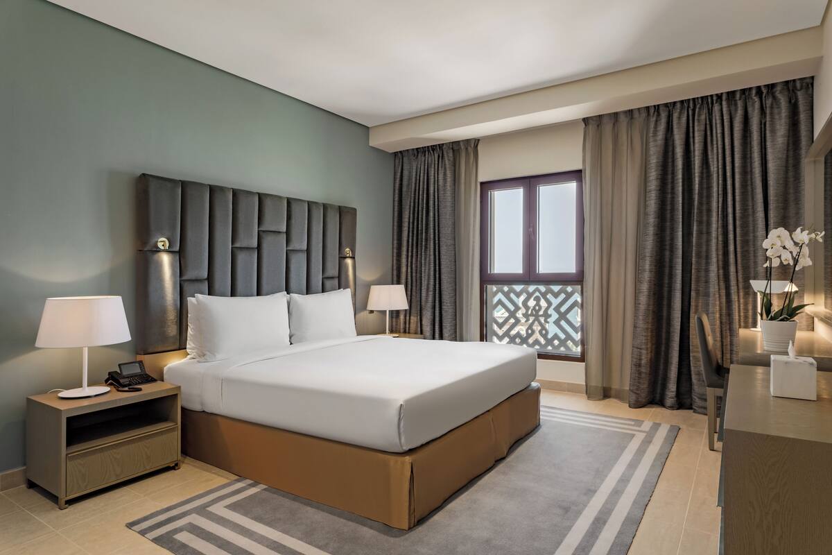 A modern hotel room with a large bed, bedside tables with lamps, a nightstand, a view of the outside through a window with curtains, and a decorative plant.