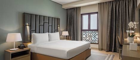 A modern hotel room with a large bed, bedside tables with lamps, a nightstand, a view of the outside through a window with curtains, and a decorative plant.
