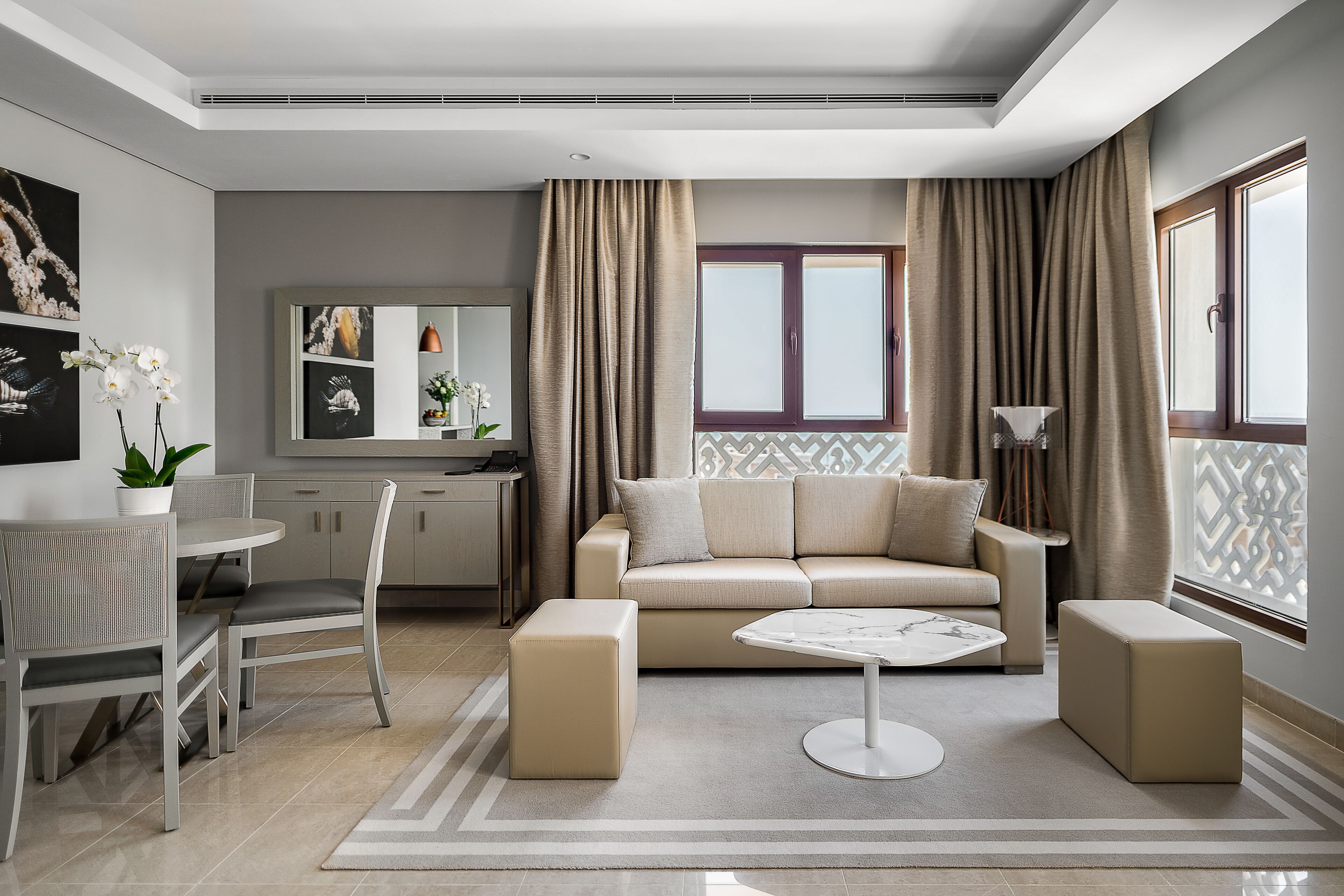 A modern living room with a beige sofa, two ottomans, a round coffee table, and a dining area with a white table and chairs.