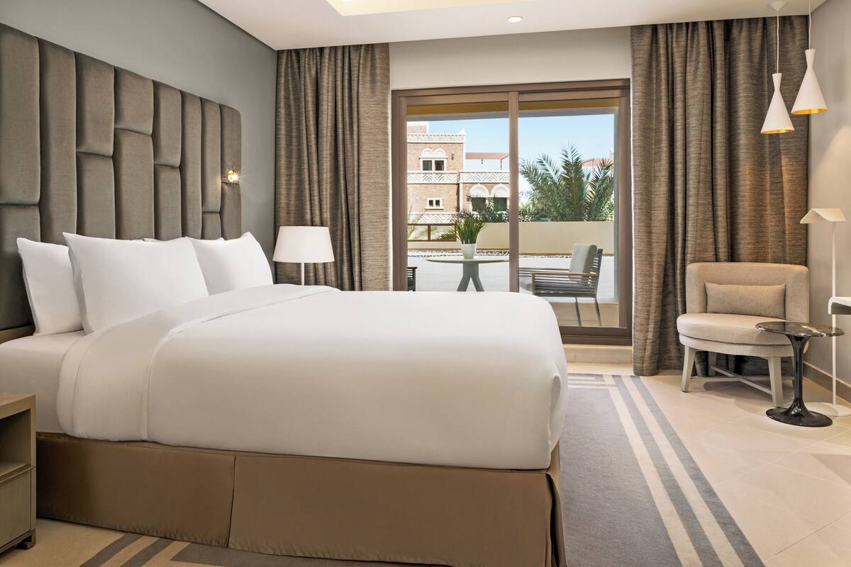 A hotel room with a large bed, a desk, a chair, and a view of the outside.